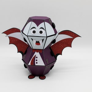 May include: A purple and white papercraft vampire with red glitter wings. The vampire has a white face with black eyes and a red mouth with fangs.