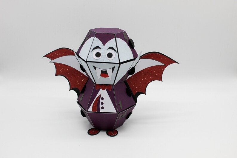 May include: A purple and white papercraft vampire with red glitter wings. The vampire has a white face with black eyes and a red mouth with fangs.