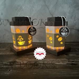 Könnte beinhalten: Zwei braune und weiße Papier-Lampen in Form einer Kaffeetasse mit grünem Band und Kaffeekorn-Designs. Die Lichter sind an und leuchten.