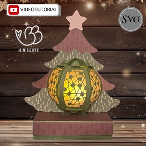 Könnte beinhalten: Ein Weihnachtsbaum aus Papier mit einer leuchtenden Laterne in der Mitte. Der Baum ist mit einem rosa Stern oben und einer braunen Basis verziert. Die Laterne ist aus Papier und hat ein geometrisches Design.