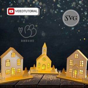 Puede incluir: Tres casas de papel blanco con techos y ventanas amarillas, iluminadas desde el interior, sobre una superficie de madera. Las casas están decoradas con pequeñas vallas blancas y árboles. El texto "VIDEO TUTORIAL" y "SVG" es visible en el fondo.