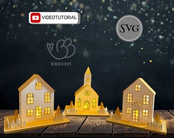 May include: Three white paper houses with yellow roofs and windows, lit from within, on a wooden surface. The houses are decorated with small white picket fences and trees. The text "VIDEO TUTORIAL" and "SVG" are visible in the background.