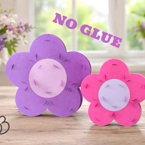Könnte beinhalten: Zwei blütenförmige Bastelarbeiten, eine lila und eine rosa, stehen auf einer Holzoberfläche. Jede hat eine hellviolette Mitte und blütenblattartige Ränder. Der Text "NO GLUE" ist oben aufgedruckt. Im Hintergrund ein Blumenarrangement.