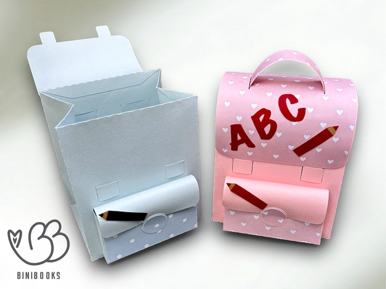 Puede incluir: Dos mochilas de papel, una azul claro con lunares blancos y otra rosa con corazones blancos. Ambas tienen un l&aacute;piz y un bolsillo con solapa en la parte delantera. La mochila rosa tiene las letras "ABC" en la parte delantera.
