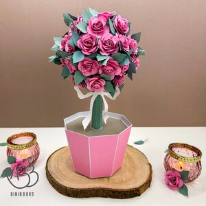 May include: A pink paper rose bush with green leaves in a pink hexagonal pot. The pot is sitting on a wooden surface. There are two small glass candle holders with pink roses on the surface.