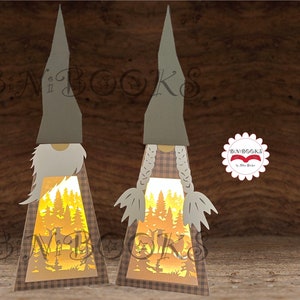 Könnte beinhalten: Zwei graue und weiße gnomenförmige Papierlaternen mit einer Waldlandschaft im Inneren. Die Laternen sind mit einem warmen Schein beleuchtet. Die Laternen stehen auf einer Holzoberfläche.