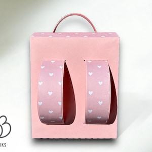 Puede incluir: Una caja de cart&oacute;n rosa con forma de mochila con un asa en la parte superior. La caja tiene dos secciones recortadas en la parte delantera, cada una con un inserto de papel con estampado de corazones rosa y blanco.