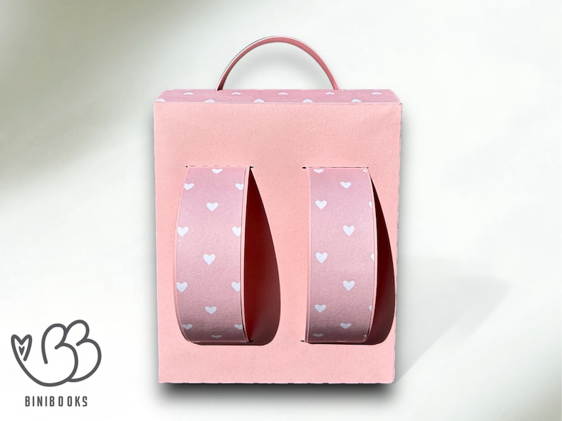 Puede incluir: Una caja de cart&oacute;n rosa con forma de mochila con un asa en la parte superior. La caja tiene dos secciones recortadas en la parte delantera, cada una con un inserto de papel con estampado de corazones rosa y blanco.