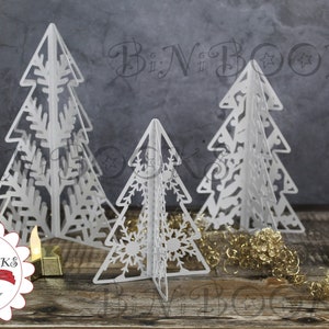 Puede incluir: Tres árboles de Navidad de papel blanco con intrincados diseños recortados. Los árboles están dispuestos sobre una superficie de madera con purpurina dorada y pequeños portavelas.