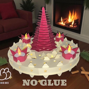 Peut inclure: Centre de table festif en papier, avec un sapin de Noël magenta à plusieurs niveaux et quatre bougeoirs avec des lumières LED. La base blanche est ornée d'étoiles. Le texte "NO GLUE" est visible.