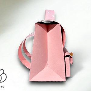 Puede incluir: Una bolsa de papel rosa con un patr&oacute;n de coraz&oacute;n blanco en el asa. La bolsa est&aacute; doblada y tiene un asa que est&aacute; doblada sobre la parte superior. La bolsa est&aacute; sentada sobre una superficie blanca.