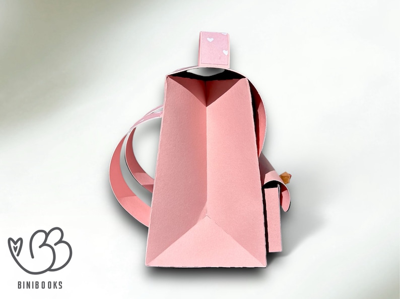 Puede incluir: Una bolsa de papel rosa con un patr&oacute;n de coraz&oacute;n blanco en el asa. La bolsa est&aacute; doblada y tiene un asa que est&aacute; doblada sobre la parte superior. La bolsa est&aacute; sentada sobre una superficie blanca.