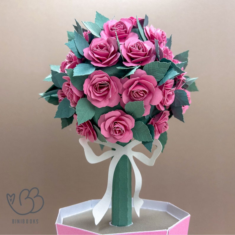 May include: A papercraft rose bouquet in a pink pot with a white bow. The bouquet is made of pink paper roses and green paper leaves.