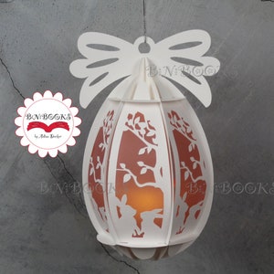 May include: White papercraft Easter egg lantern with a bow at the top. The egg is decorated with a tree and bunnies. The lantern is lit from within, casting a warm glow.