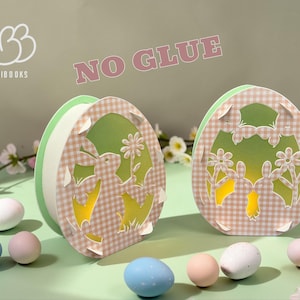 May include: Two Easter egg-shaped paper cutouts with bunny and flower designs, set against a green backdrop. The cutouts have a gingham pattern and are surrounded by pastel-colored eggs and flowers. The text "NO GLUE" is visible.