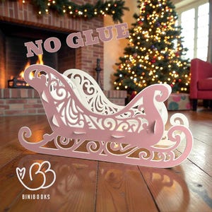 May include: A decorative, laser-cut sleigh in pink and white, with intricate scrollwork. The sleigh is made of wood and is displayed on a wooden floor. The text "NO GLUE" is visible above the sleigh. The sleigh is a Christmas decoration.