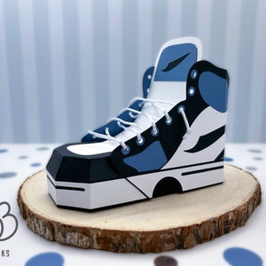 May include: A papercraft model of a blue and white high-top trainer with black accents. The trainer is sitting on a wooden base.