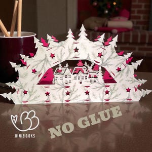 May include: A white and pink Christmas village paper cut-out with fir trees and stars. The cut-out features a village scene with houses and trees. The words "NO GLUE" are printed on the bottom. A burgundy mug with wooden sticks is in the background.