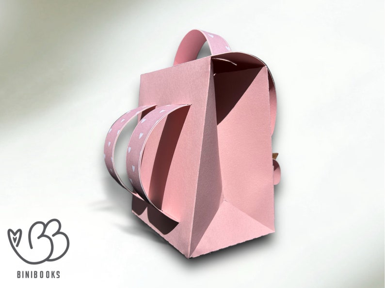 Puede incluir: Una bolsa de regalo de papel rosa con un patr&oacute;n de corazones blancos en las asas. La bolsa tiene un asa que se pliega sobre la parte superior.