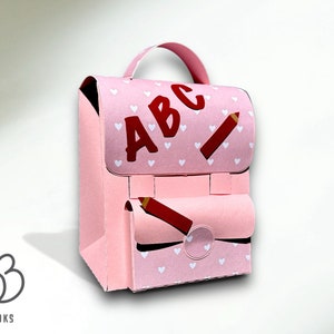 Puede incluir: Una mochila de cart&oacute;n rosa con un patr&oacute;n de corazones blancos y las letras "ABC" en rojo. La mochila tiene un dise&ntilde;o de l&aacute;piz rojo en la parte delantera y un bolsillo con un dise&ntilde;o de l&aacute;piz rojo.