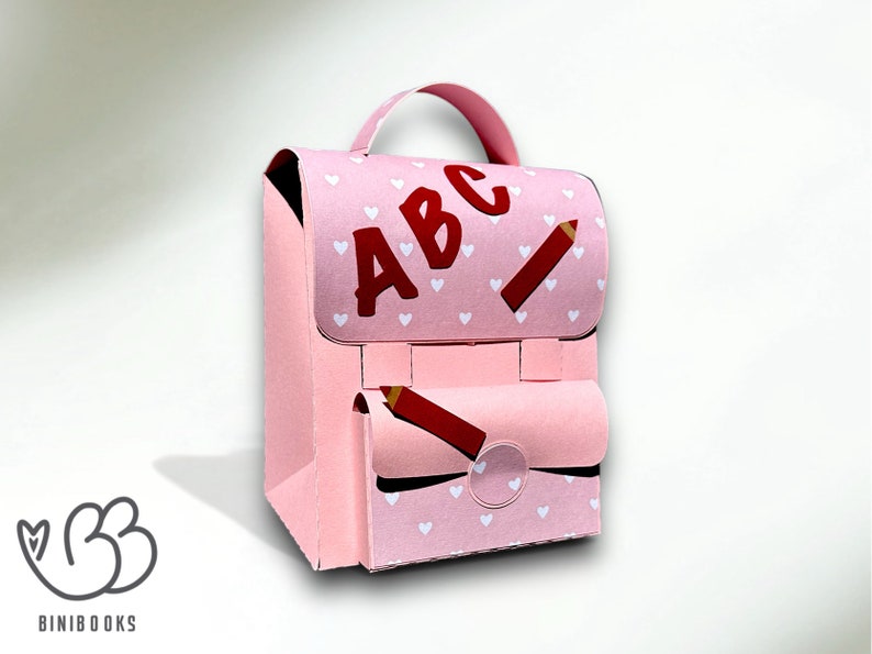 Puede incluir: Una mochila de cart&oacute;n rosa con un patr&oacute;n de corazones blancos y las letras "ABC" en rojo. La mochila tiene un dise&ntilde;o de l&aacute;piz rojo en la parte delantera y un bolsillo con un dise&ntilde;o de l&aacute;piz rojo.