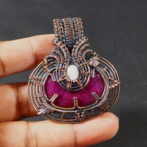 May include: A handcrafted pendant with a wire-wrapped design. The pendant features a vibrant magenta stone at the center, accented by a small, oval, white stone. The intricate wirework is a mix of copper and black tones, creating a unique, artistic piece.