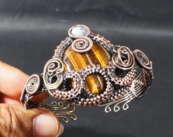 May include: A handcrafted copper wire bracelet featuring a large, oval tiger's eye stone. The bracelet has intricate wirework with spiral and woven designs, and a small, round white stone accent.