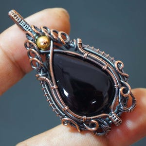 Może przedstawiać: Czarny kamień szlachetny zawieszony na srebrnym i miedzianym drucie. Zawieszka ma małą złotą kulkę.