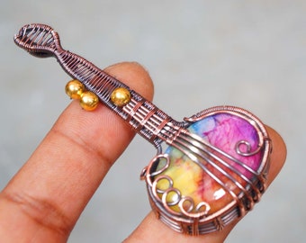Puede incluir: Un colgante de alambre envuelto de colores con forma de guitarra con una piedra de color arcoíris en el centro. El colgante está hecho de alambre de cobre y tiene cuentas doradas en el cuello.
