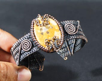 May include: A handcrafted wire-wrapped cuff bracelet. The bracelet features a large, oval-shaped, yellow and brown stone at the center. The cuff is made of woven silver and copper wire with decorative spiral accents. The bracelet is approximately 2 inches wide.