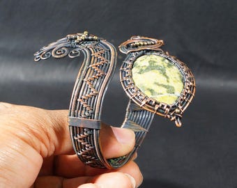 May include: Handmade copper wire cuff bracelet with a large oval green and yellow stone. The bracelet features intricate wire weaving and decorative swirls. The metal has a dark, aged finish, giving it a rustic appearance.