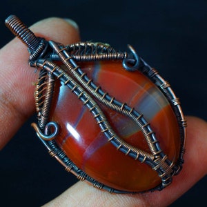 May include: A red agate gemstone pendant with copper wire wrapping. The wire is wrapped in a decorative pattern around the stone, creating a unique and intricate design.
