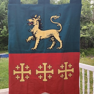 Könnte beinhalten: Ein rechteckiges Stoffbanner, das an einer Holzstange hängt, mit einem blaugrünen oberen Teil mit einer goldenen Löwenillustration und einem roten unteren Teil, der drei goldene Kreuzsymbole mit jeweils vier kleinen Kreuzen zeigt.