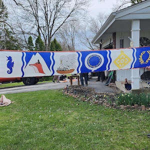 May include: A long, rectangular banner with a red border and a white central panel. The banner features various heraldic symbols in blue, gold, and red, including a seahorse, fox, pelican, chain, and laurel wreath. The banner is held up by two people.