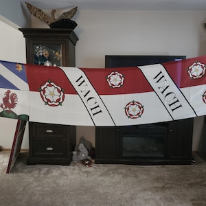 May include: A long flag with a design featuring a red rooster, stars, and a diagonal stripe with the word "WACH". The flag has a white background with red and white stripes and floral emblems.