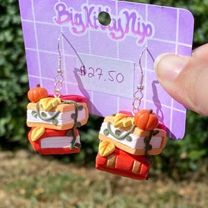 May include: Handmade earrings with a book theme, featuring miniature stacks of books in autumnal colours. Each earring has a small pumpkin and decorative vines. The earrings are suspended from silver-coloured hooks. The earrings are on a purple card.