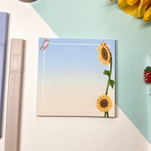 May include: A blue and white gradient notepad with a sunflower and butterfly design. The notepad has a white border and is perfect for writing notes or reminders.