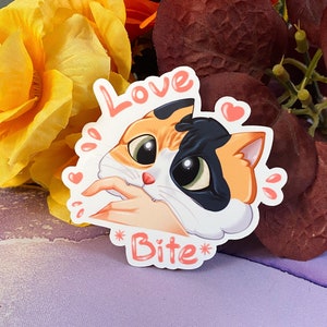 May include: A sticker featuring a cartoon calico cat with big eyes and a pink nose. The cat is biting a finger and the sticker says "Love Bite" in pink lettering.