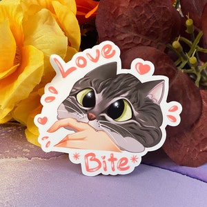 May include: A white sticker with a cartoon gray and white cat with big eyes. The cat is biting a finger. The sticker says "Love Bite" with hearts around the cat.