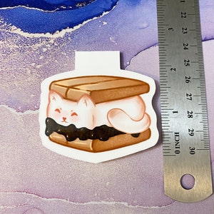 May include: A white cat-shaped marshmallow is sandwiched between two graham crackers with chocolate spread. The marshmallow has a happy expression with closed eyes and a small pink nose.