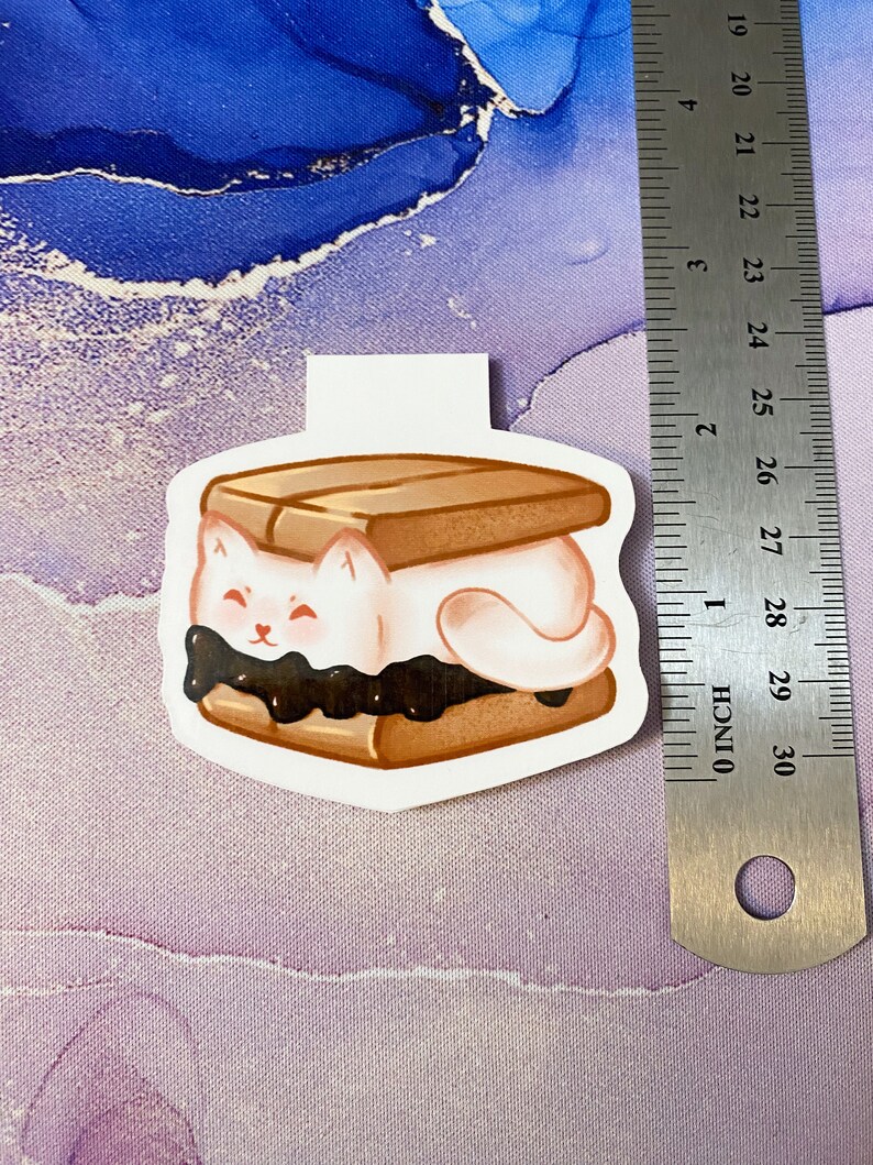 May include: A white cat-shaped marshmallow is sandwiched between two graham crackers with chocolate spread. The marshmallow has a happy expression with closed eyes and a small pink nose.