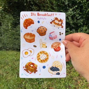 May include: A sheet of stickers featuring various breakfast foods, including pancakes, waffles, croissants, muffins, eggs, and biscuits. The stickers are illustrated in a cartoon style with bright colors and whimsical details. The text "It's Breakfast!" and "BigKittyFlip" are printed on the sheet.