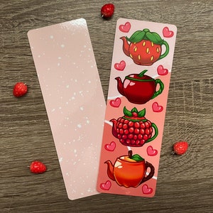 May include: Two rectangular bookmarks. One is light pink with white speckles. The other has four illustrated teapots: a strawberry, a raspberry, and an apple teapot, with pink hearts. The teapots are red, orange, and pink.