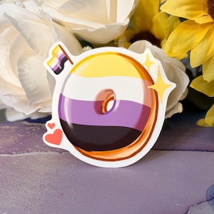 May include: A sticker featuring a donut design with the non-binary pride flag colors: yellow, white, purple, and black. The donut has a brown outline and is decorated with two hearts and starbursts. The sticker is placed in front of white roses and yellow sunflowers.