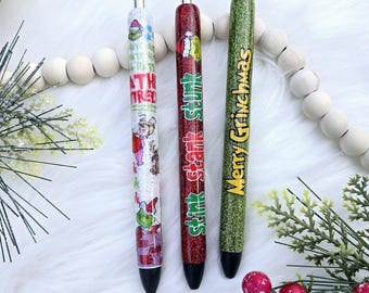 May include: Three glitter pens with Christmas-themed designs. The pens feature the text "Stink Stank Stunk", "Merry Grinchmas", and "The Grinch" with illustrations of the Grinch.