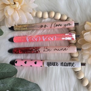 May include: Four glitter pens with black tips and silver accents. The pens are decorated with pink, gold, and red glitter. Each pen has a different message: "p.s. I love you", "Kisses", "Be mine", and "your name".