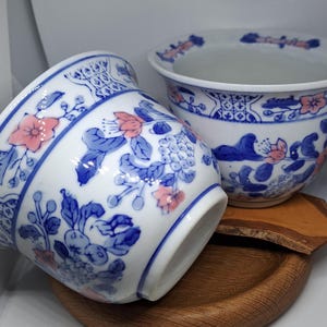 May include: Two white ceramic plant pots with blue and pink floral patterns. The pots have a wide rim and a rounded body. The floral design includes stylized flowers and leaves. The pots are on a wooden surface.