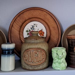 May include: A selection of vintage kitchen items on a shelf. There are mugs with fruit and cartoon designs, a sugar jar, a salt shaker, a wooden plate, and decorative ceramic pieces. The items are in various colours, including brown, green, and white.
