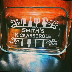 Può includere: Pirofila rettangolare in vetro trasparente con testo inciso e disegni di utensili da cucina. Il testo recita "SMITH'S KICKASSEROLE". Il piatto ha angoli arrotondati e manici.