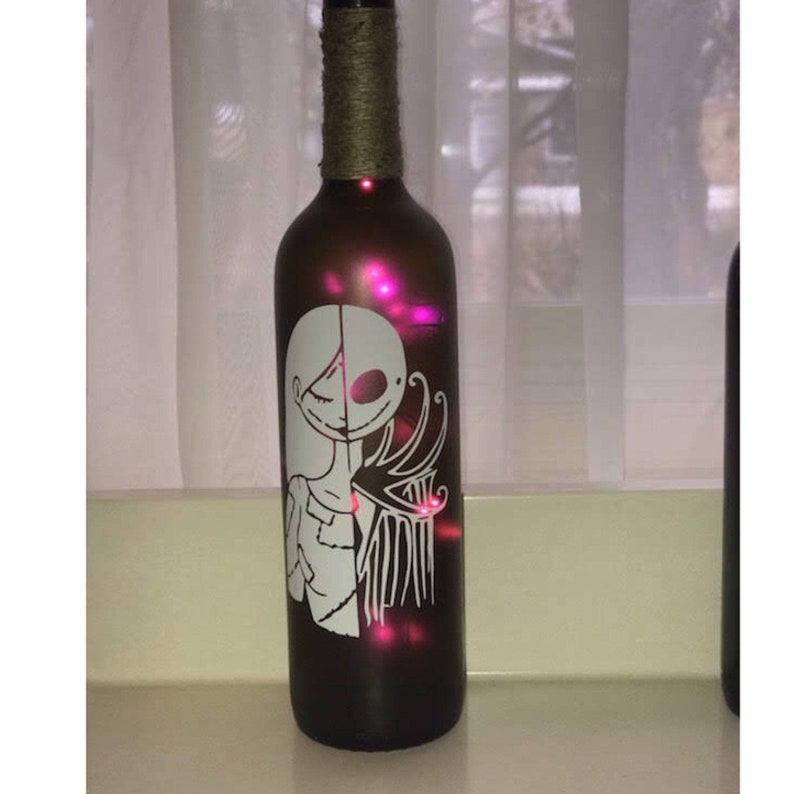May include: A brown glass wine bottle with a white silhouette of Jack Skellington and Sally from the Nightmare Before Christmas. The bottle is decorated with twine and has a pink glow from inside.